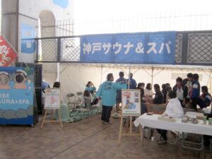 青い神戸サウナ＆スパの名前が掲示され、多くの人が足湯とマッサージを受けている。